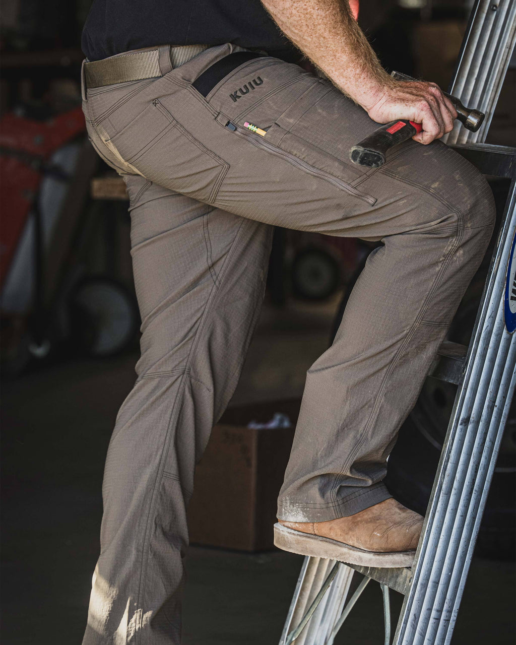 Man stepping up onto a metal ladder while wearing Workgrade Pant in Rodeo Brown, and a hand holding a hammer in a workshop setting.