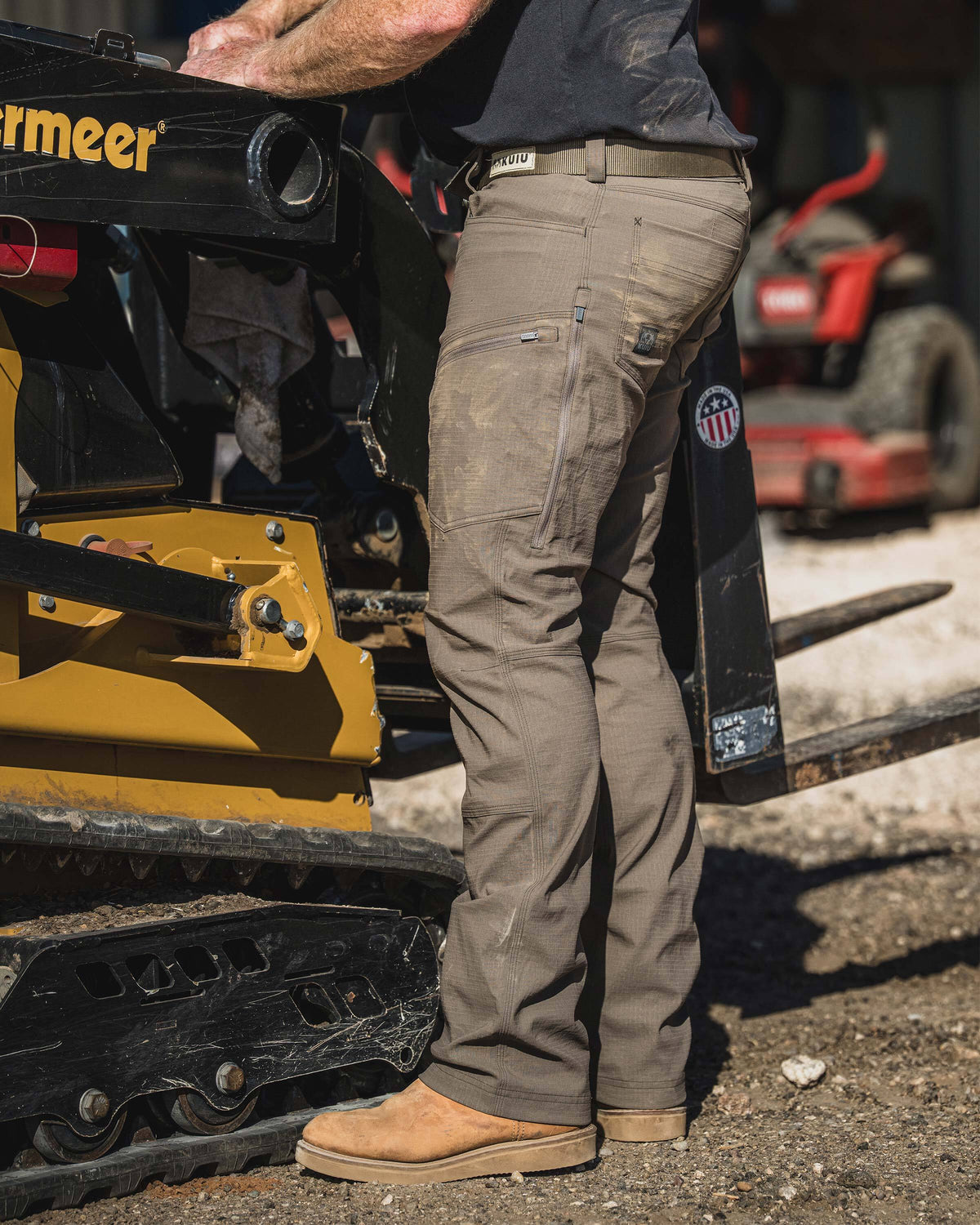 Side view of a man standing on gravel beside tracked construction equipment in an outdoor jobsite setting, wearing Motive Workgrade Pant in Rodeo Brown
